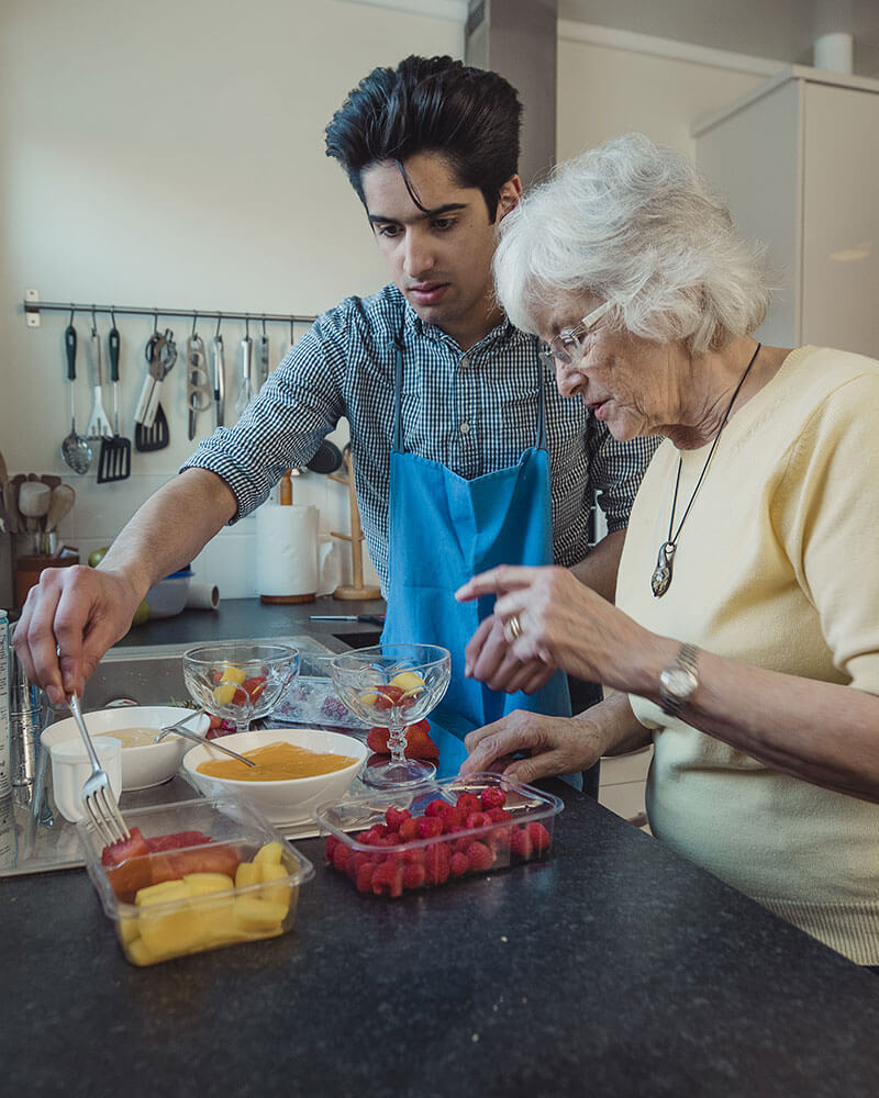 Young man and elderly woman putting together fruit cups at the kitchen counter